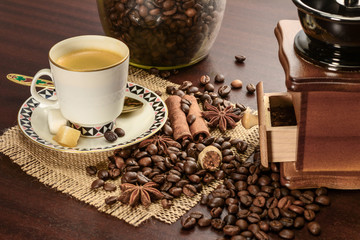 Cup of coffeee with saucer on a jute napkin. Vintage still life on wooden table with sugar, anise, cinnamon, grinder and coffee jar