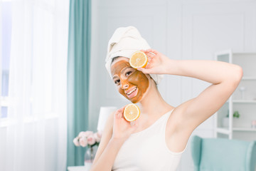 Beauty procedures skin care concept. Young woman with brown facial mud clay mask on her face in bathroom, with white towel on head, holding lemon halfs