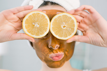 Young pretty girl with brown mud mask on face holding lemon fruit halves, covering eyes. Teen girl taking care of her skin. Beauty treatment.