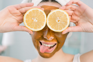 Young pretty girl with brown mud mask on face holding lemon fruit halves, covering eyes. Teen girl taking care of her skin. Beauty treatment.