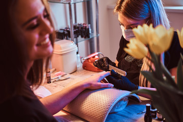 Nail master is working on client nails with nail file. Master is wearing protection mask and black latex gloves.