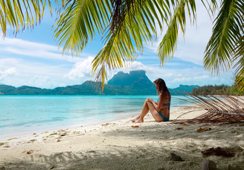 Young female tourist in blue bikini sits on sandy beach and observes the ocean.