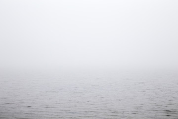 Misty morning over the lake. Gray thick fog over the water surface closes the horizon. Dull day.