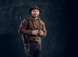 Mature hunter with rifle holding binoculars and looking at a camera. Studio photo against dark wall...