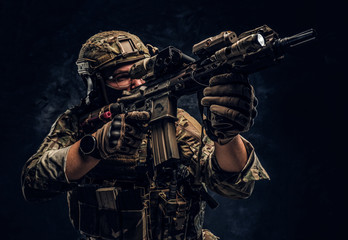 Fototapeta na wymiar Close-up studio photo against a dark wall. The elite unit, special forces soldier in camouflage uniform holding an assault rifle with a laser sight and aims at the targe