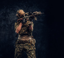 The elite unit, special forces soldier in camouflage uniform holding an assault rifle with a laser sight and aims at the target. Studio photo against a dark wall