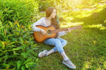 Young hipster woman sitting in grass and playing guitar on park or garden background. Teen girl...