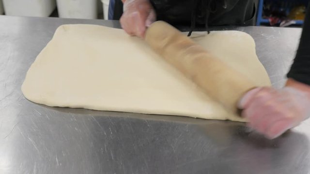 Woman Uses Wood Rolling Pin To Flatten Rectangle Of Dough On Metal Table. Baker uses wooden rolling pin to evenly flatten dough.