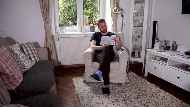 Young casual man reading a book in his home sitting in an arm chair