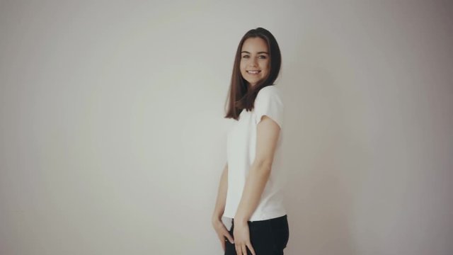 Young beautiful girl posing in front of the camera in white cotton blank t-shirt and black jeans with white isolated wall on the background, mock up of clothes, happy smiling and laughing