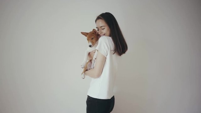 Cute young hipster girl holding a lovely basenji puppy dog, hugging and kissing each other. Girl wearing blank white cotton t-shirt standing in front of white wall. Love between pet and his owner