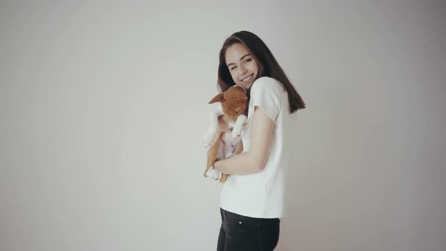 Pretty teenage girl playing with her basenji dog at home, hugging him and kissing. Little cute puppy dog is sleepy and the girl is laughing very happy. Isolated white wall on the background.