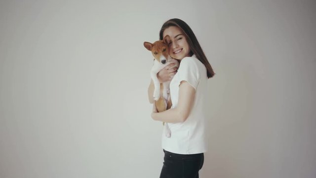 Cute young hipster girl holding a lovely basenji puppy dog, hugging and kissing each other. Girl wearing blank white cotton t-shirt standing in front of white wall. Love between pet and his owner