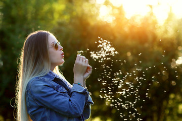 Young spring fashion woman blowing dandelion in spring garden. Springtime. Trendy girl at sunset in spring landscape background. Allergic to pollen of flowers. Spring allergy.