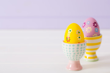 Two colorful painted Easter eggs in vibrant modern egg stands on pastel lilac background.