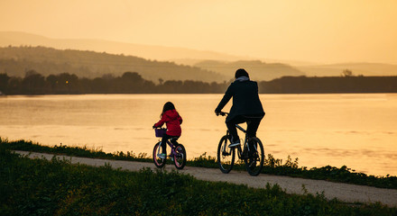 Father and child riding bicycles by the river in sunset