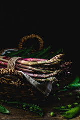 Fresh green and purple asparagus, green peas in wicker basket, hard light, deep shadows, dark background - gourmet organic food