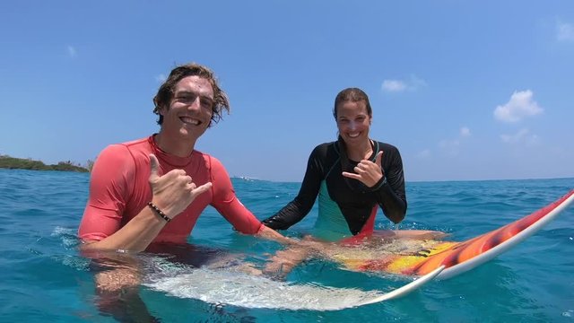 Happy couple of surfers sit on their surfboards in the tropical water and they both show the Shaka sign
