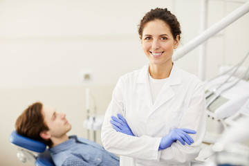 Waist up portrait of female dentist smiling at camera while consulting patient in background, copy...