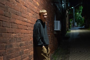 Serious looking teenager leaning against a brick wall in an alleyway at night.