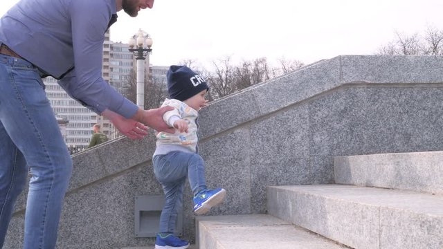 bearded man in sunglasses holding his little son by the hand helps the baby boy to climb the stairs in the city park on sunny spring day background in slow motion middle shot 4K video