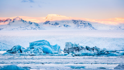 Icebergs float on Jokulsarlon glacier lagoon at sunrise, with background mountain peaks lit by sunrise, in Iceland.
