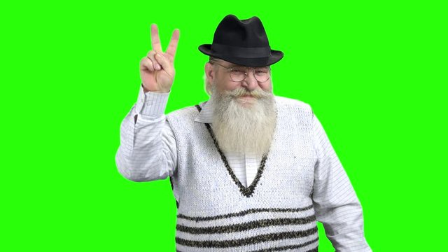 Cheerful senior man demonstrating victory sign. Elderly bearded man showing peace gesture while standing on green screen background.