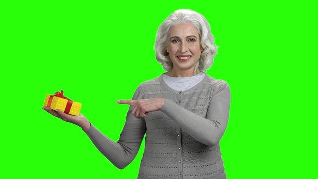 Happy elderly woman showing gift box. Senior smiling woman pointing with finger on gift box. Green screen chroma key background for keying.