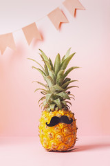 Funny dressed up pineapple with a black mustache pink pastel background with festive flags. Minimalism. Vertical frame.