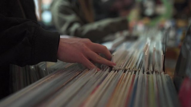 Slow motion shot of a male hand flipping through a stack of vinyl records in a record store. There are rows of records in the background, shallow depth of field, gritty with slight light flicker.