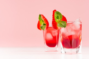 Red alcoholic shots of sweet fruit liquor with ice cubes, strawberry slice, green mint on pastel soft light pink background, copy space.