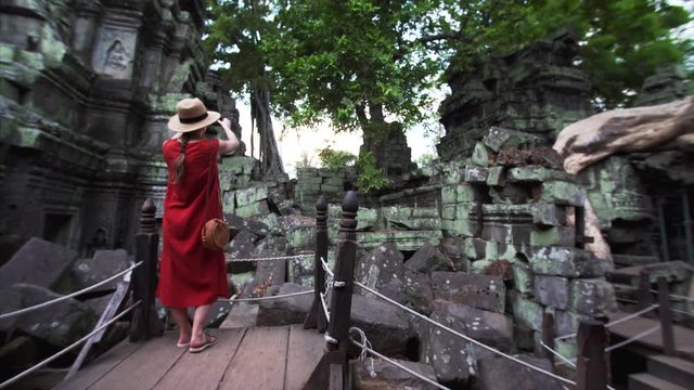 Woman in red dress walks in the frame to take picture of picturesque ruins and towers of famous Ta Prohm temple (Angkor Wat complex) in Cambodia. Beautiful sunny day