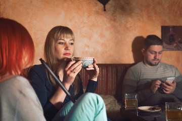 Smoke hookah shisha in bar and nightclub, team of friends two girls and man, have fun, drink tea. a man alone uses a smartphone, uses the network, Wi-Fi