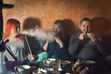 Smoke hookah shisha in bar and nightclub, team of friends two girls and man, have fun, drink tea.