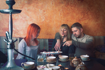 Smoke hookah shisha in bar and nightclub, team of friends two girls and man, have fun, drink tea. eat oriental sweets, Turkish delight.