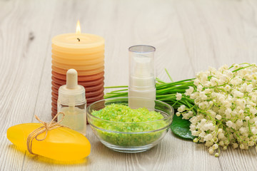 Bottles with cream for face skin and aromatic oil, bowl with sea salt, burning candle, soap and bouquet of lilies of the valley.