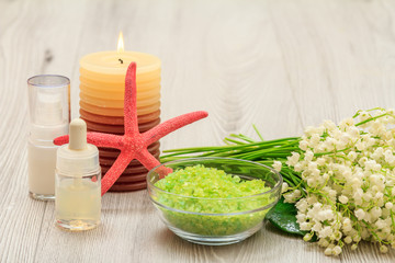 Bottles with cream for face skin and aromatic oil, bowl with sea salt, burning candle, starfish, hand massager and bouquet of lilies of the valley.