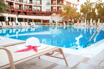 Summer hat lying on deck chair in front of swimming pool.