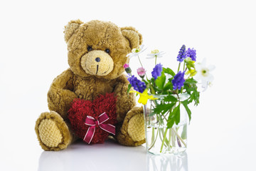 Ein Teddybär mit Herz neben einer Vase mit Blumen.