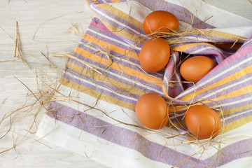 Five brown chicken eggs and dry grass on a striped towel on a beige wood texture background