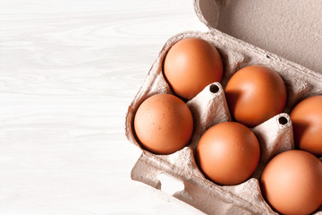 Six brown chicken eggs lie in a cardboard box located diagonally on a wood texture background. Top...