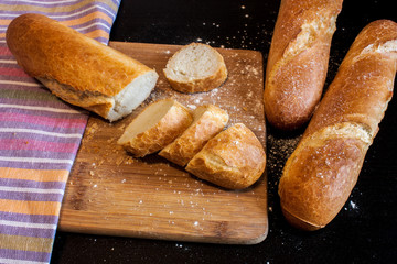 Sliced fresh crusty baguette and two whole baguette on a wooden board against the background of striped towels on a dark table