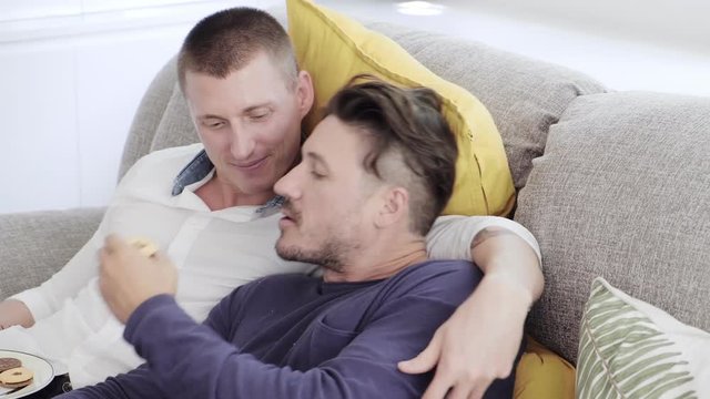 Young gay male couple relaxing on couch in living room. Caucasian, european men sitting together on sofa. Watching tv and holding cookie plate. Happy gay friendship, relationship, lifestyle concept. 