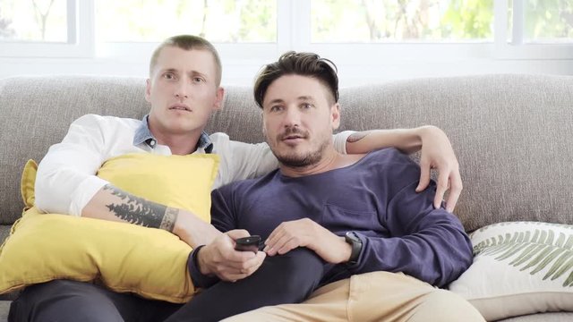 Young gay male couple relaxing on couch in living room. Caucasian, european men sitting together on sofa. Enjoy watching tv, tense mood. Happy gay friendship, relationship, lifestyle concept. 