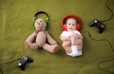 Adorable baby boy with teddy bear, headphones and joypads lying on soft plaid