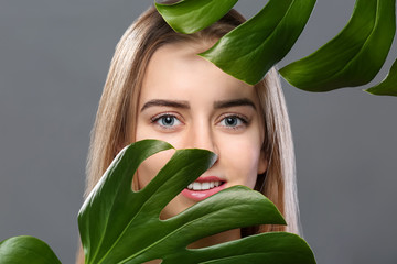 Beautiful young woman with laminated eyelashes and tropical leaves on grey background