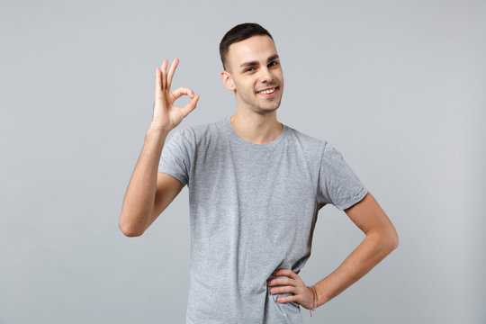 Portrait of attractive young man in casual clothes standing, looking camera and showing OK gesture isolated on grey wall background. People sincere emotions, lifestyle concept. Mock up copy space.