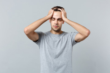 Portrait of tired young man in casual clothes keeping eyes closed, putting hands on head isolated on grey wall background in studio. People sincere emotions, lifestyle concept. Mock up copy space.