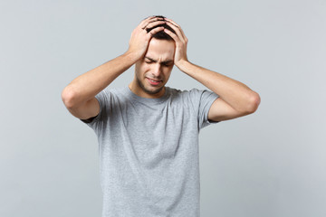 Fototapeta premium Portrait of crying dissatisfied young man in casual clothes keeping eyes closed putting hands on head isolated on grey wall background. People sincere emotions, lifestyle concept. Mock up copy space.