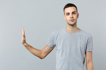 Indifferent young man in casual clothes looking camera, showing stop gesture with palm aside isolated on grey wall background in studio. People sincere emotions, lifestyle concept. Mock up copy space.